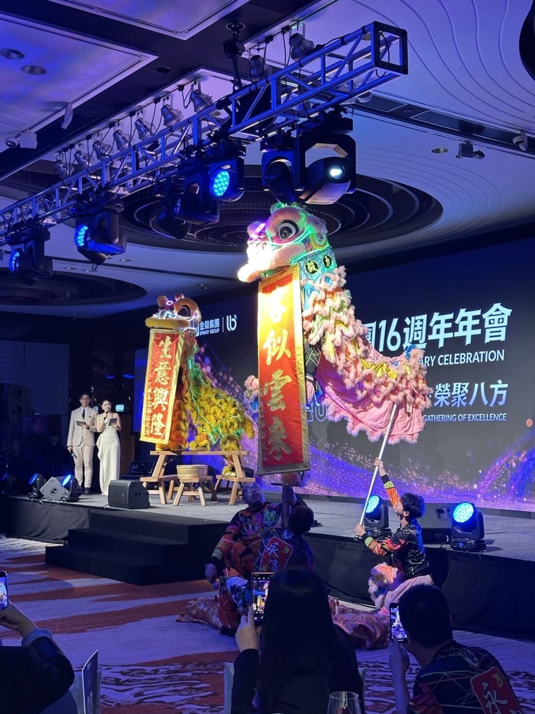 公司周年春茗晚宴：兩隻醒獅站在舞台前展開賀年揮春，司儀在台上，賓客舉起手機拍攝