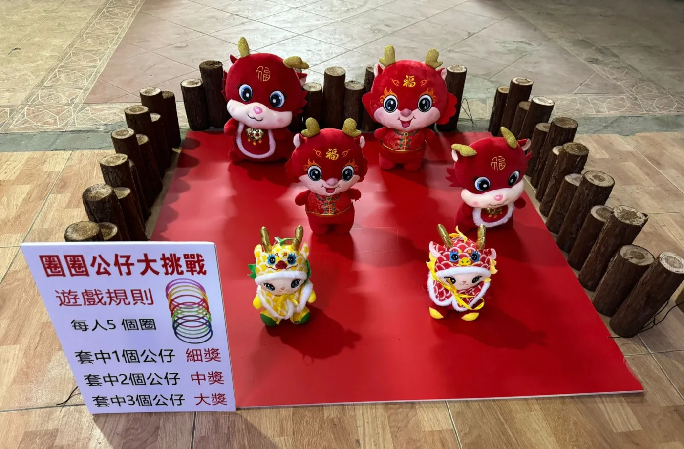 圈圈公仔攤位實景：紅色地墊上放著七隻龍年公仔，四周以木樁圍邊，前方告示寫明每人 5 個圈，套中 1 個公仔得細獎、2 個得中獎、3 個得大獎